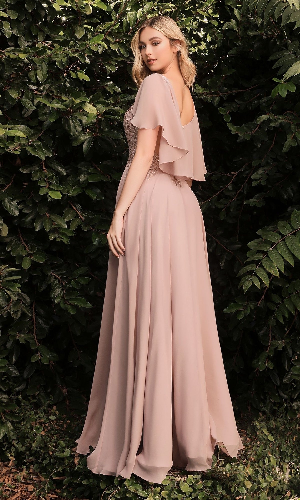 A woman stands outdoors before lush greenery, wearing the Ladivine HT101 Cape-Sleeve Long A-Line Prom Dress in light mauve chiffon with beaded details, turning over her shoulder.